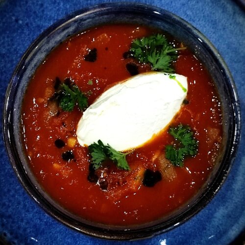 Zupa pomidorowa w niebieskiej misce na talerzu, z kleksem śmietany i świeżą pietruszką, podana na drewnianym stole.