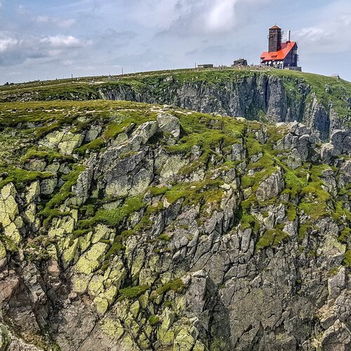 Czerwony budynek na skalistej górze z zieloną roślinnością pod błękitnym niebem.