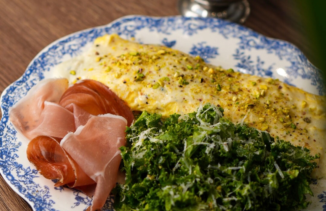 Kremowy omlet z pistacjami, cienkie plastry prosciutto i jarmuż z parmezanem na niebieskim talerzu w kwiatowy wzór