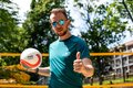 Man in sunglasses holding a volleyball and giving a thumbs-up on a hotel outdoor court with balconies and trees
