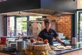 Chef in open kitchen plating desserts on counter with rustic wooden display, fresh apples, stacked plates and brick wall.