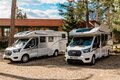 Two modern white motorhomes parked by a tiled-roof lodge amid pine trees, suggesting self-catering rentals for a peaceful forest getaway