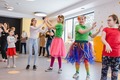 Family event room with children dancing and a costumed entertainer, balloons, parents watching and a dessert table near large windows.