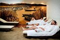 Hotel spa relaxation room with sand floor, cushioned loungers and three guests in white robes resting under a warm lakeside mural.