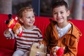 Two smiling children sitting on a red sofa, holding plush toy firefighters and a small gift box, playful family moment.