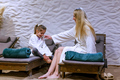 Woman and young girl in white robes share a playful moment on spa loungers with teal towels, textured wall and pampas grass