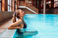 Guest relaxing in an indoor hotel pool, leaning on the edge by large windows, with wooden beams and a lounge staircase.