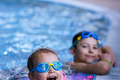 Two children in an indoor hotel pool — toddler with pink floral arm floaties and blue goggles laughing at the edge, older child leaning nearby.