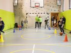 Dzieci kozłują piłki przez pachołki w sali sportowej z ścianką wspinaczkową, miękkimi ścianami i instruktorami