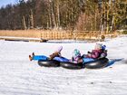 Family tubing on a snowy hill: kids in winter gear riding connected inner tubes past a wooden boardwalk and forest.