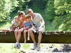 Family of four sitting on a wooden footbridge over a shallow stream, children dipping feet, sunlit forest backdrop.