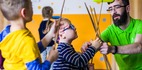 Animacje dla dzieci: instruktor pomaga maluchom kręcić kolorowymi talerzami na patykach w jasnej sali z pomarańczowymi kropkami.