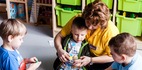 Sala zabaw: opiekunka w żółtej koszulce pomaga dzieciom siedzącym w kole sortować kolorowe koraliki przy zielonych pojemnikach.