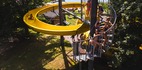 Nad jeziorem żółta spiralna zjeżdżalnia wodna, dzieci na schodach, leżaki i widok na spokojne jezioro.