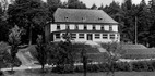 Historyczny, dwukondygnacyjny hotel z dachówką i dużymi oknami na parterze, pośród starych drzew i zadbanych trawników.