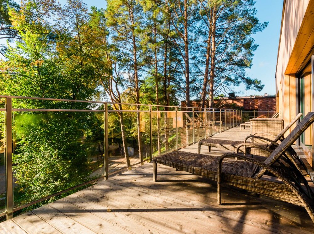 Prywatny drewniany balkon z wiklinowymi leżakami, szklaną balustradą i widokiem na korony drzew, słoneczne miejsce relaksu.