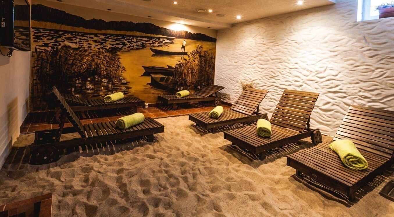 Sala relaksu spa z podłogą z piasku, drewniane leżaki z zielonymi zwiniętymi ręcznikami, ciepły mural z łodziami i trzciną.