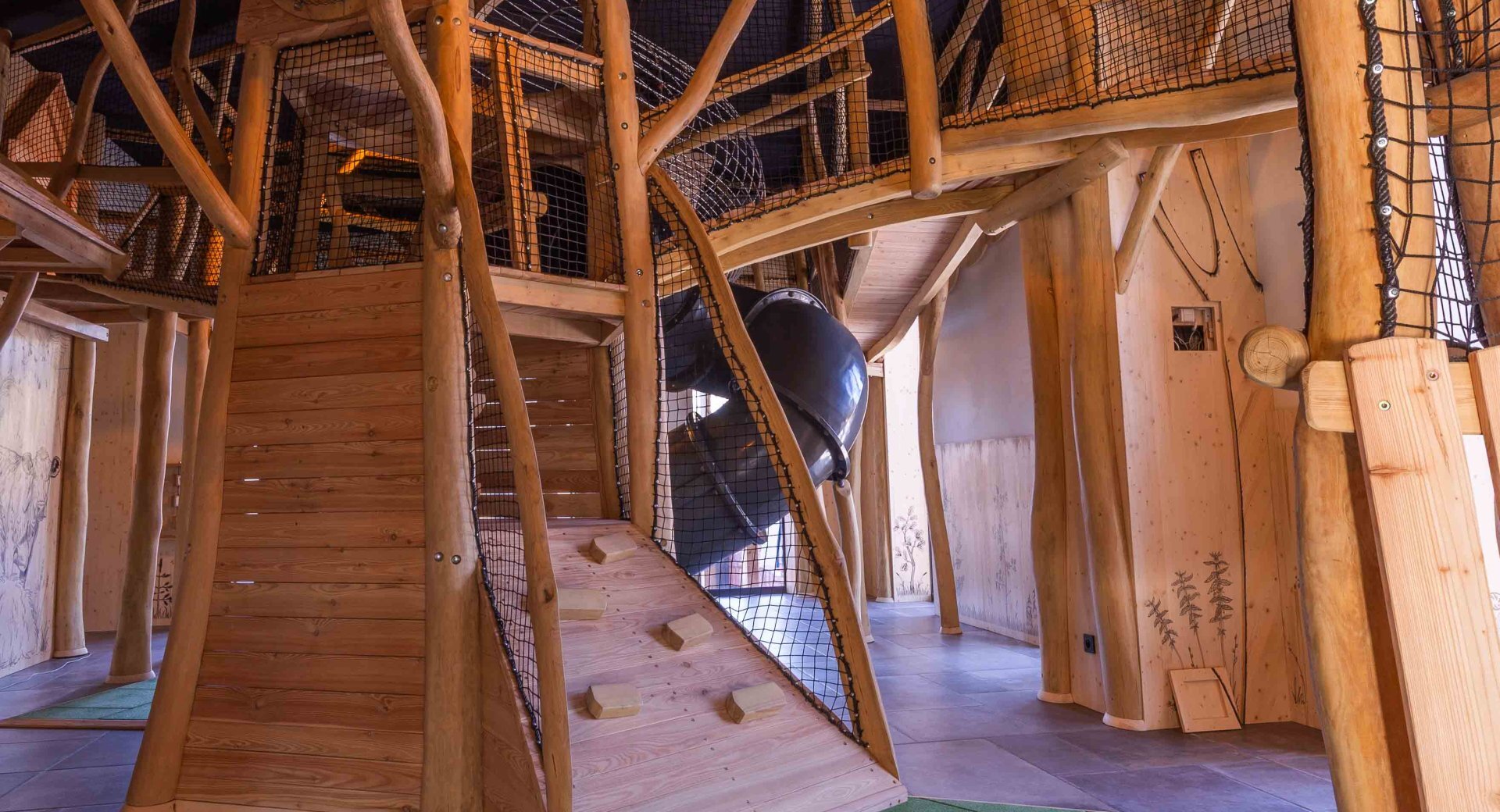 Indoor natural-wood play structure with climbing ramp, safety netting and enclosed tube slide in the hotel's family play area