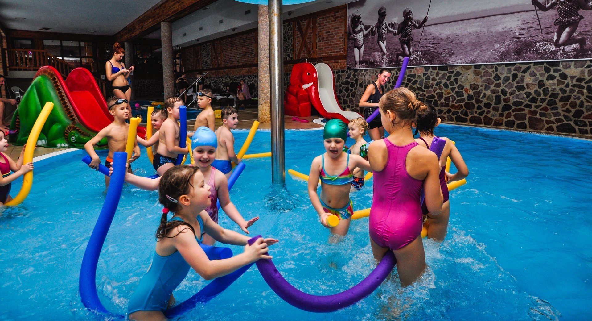 Indoor family pool with children playing with colorful foam noodles near a green dinosaur and red elephant slide, supervised by staff.