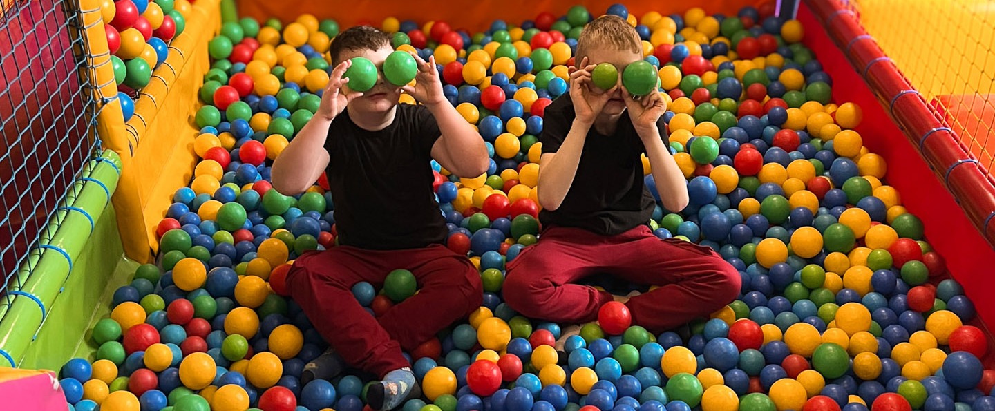 Children's play area ball pit with colorful netted soft-play structure and two children holding green balls to their eyes