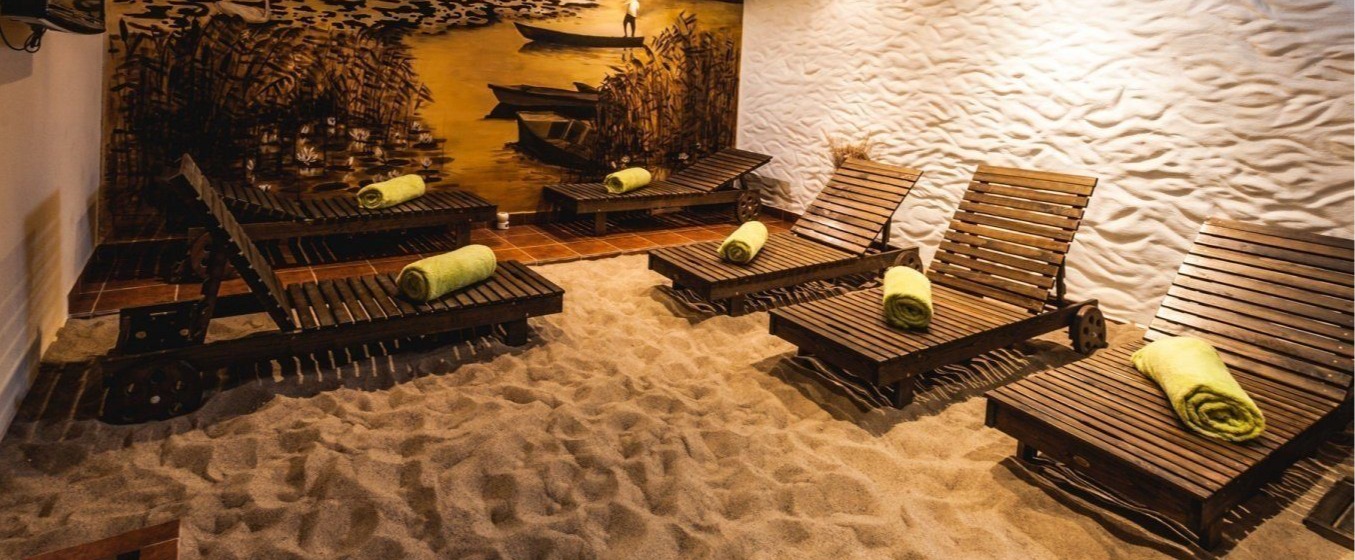 Indoor spa relaxation room with sand floor, wooden lounge chairs each with rolled green towel, warm mural of boats and reeds.