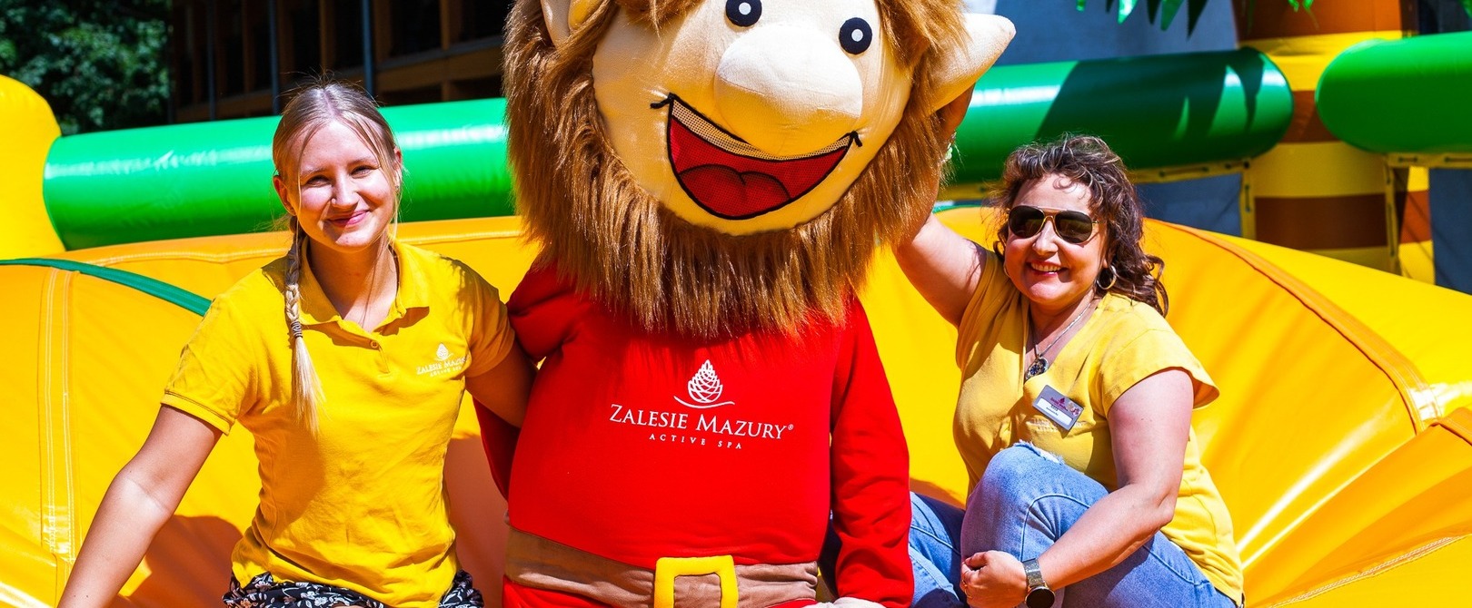Resort staff smile beside a red-costumed mascot on a bright yellow inflatable play area — family-friendly kids' entertainment at Zalesie Mazury