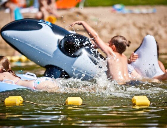Children splashing on an inflatable orca in a lakeside family swim area marked by yellow buoys