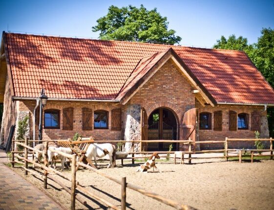 Rustic brick stable with red-tiled roof, wooden shutters and fenced sandy paddock where ponies graze.