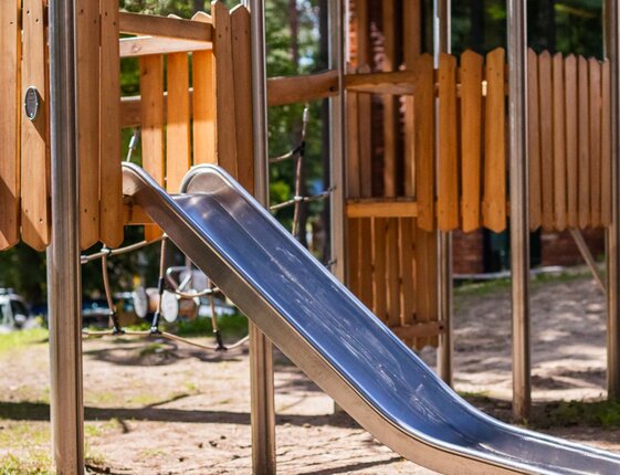 Child climbing a rope ladder to a wooden playground platform with a metal slide among trees in the hotel's family play area.