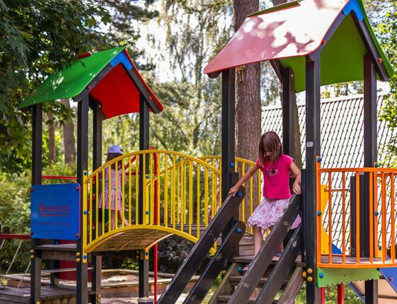 Family-friendly hotel playground with children on colorful climbing structure, bridge and shaded roofs.