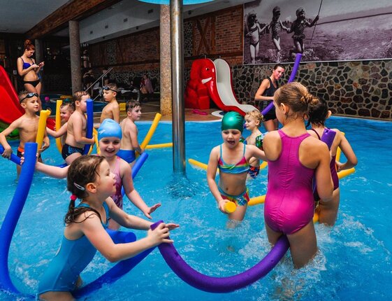 Indoor family pool with children playing with colorful foam noodles near a green dinosaur and red elephant slide, supervised by staff.