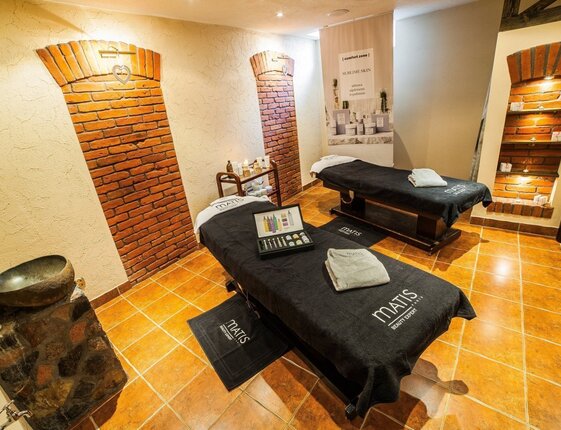 Couples spa treatment room with two massage tables, Matis towels, skincare display, warm tile floor and exposed brick arches.