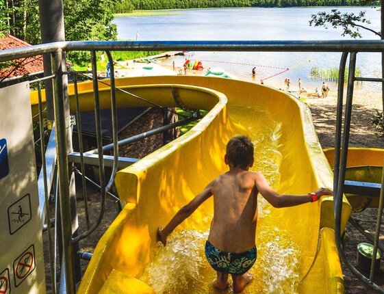 Child on yellow water slide at lakeside resort, view of sandy beach, swimmers and forested shoreline at Zalesie Mazury Active & Spa.