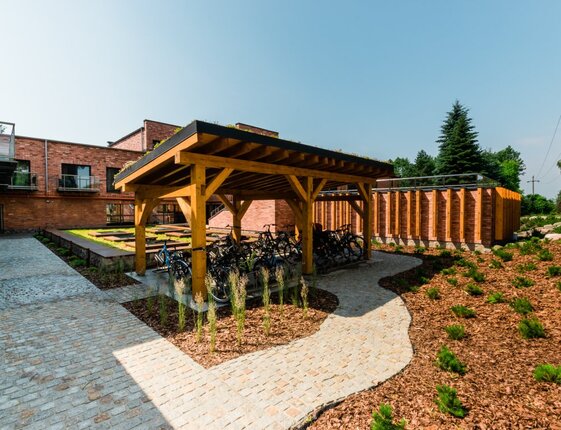 Covered wooden bike shelter with multiple bicycles, paved courtyard, brick hotel buildings and landscaped garden beds