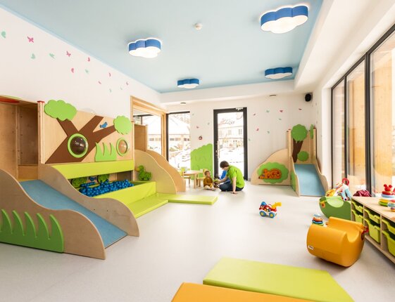 Bright hotel kids' playroom with indoor slides, ball pit, cloud lights, toys and a staff member playing with a child.