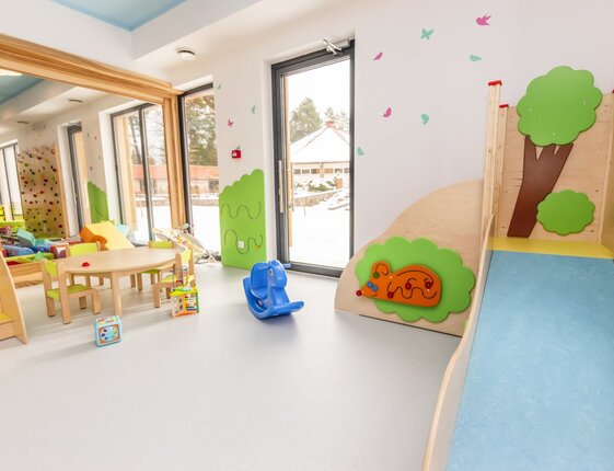Bright children's playroom with wooden slide, small table and chairs, toys, indoor climbing wall and large windows with snowy outdoor view