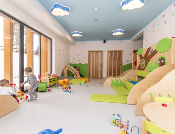 Bright hotel children's playroom with wooden slides, ball pit, floor mats, cloud lights, large windows and toddlers playing.