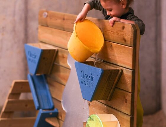 Toddler pouring water from a yellow bucket into a wooden water-play wall over indoor sand in a family-friendly kids' play area.