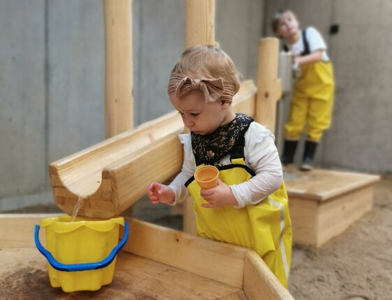 Toddler in yellow waterproof overalls and bow headband scoops water with a cup into a yellow bucket from a wooden water play table; another child behind.
