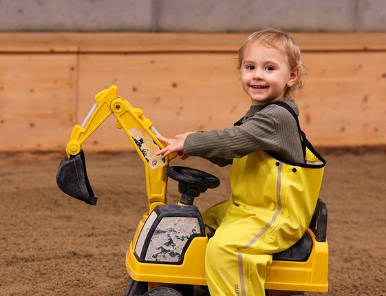 Smiling toddler in yellow rain suit driving a toy excavator in a hotel's indoor kids' play area with soft sand floor.