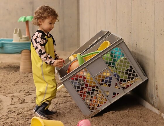 Toddler in yellow waterproof suit playing with a crate of colorful beach toys in an indoor sand play area