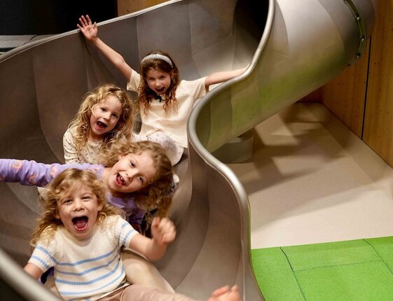 Four joyful children sliding down a wide indoor metal slide in the hotel's family playroom
