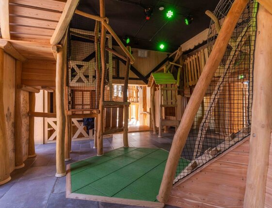 Indoor children's play area with rustic wooden treehouse frames, climbing nets, soft green safety mat and ceiling lights.