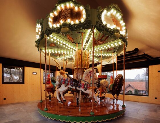 Indoor vintage carousel with hand-painted horses and animals, lit bulbs on polished wooden base in wood-paneled room with countryside view.