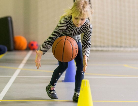 Young girl dribbling a basketball through training cones in an indoor gym, focused on the ball
