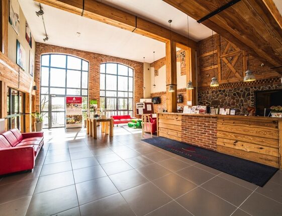 Sunlit hotel lobby with exposed brick and timber beams, large arched windows overlooking trees and water, red sofas and wooden reception desk