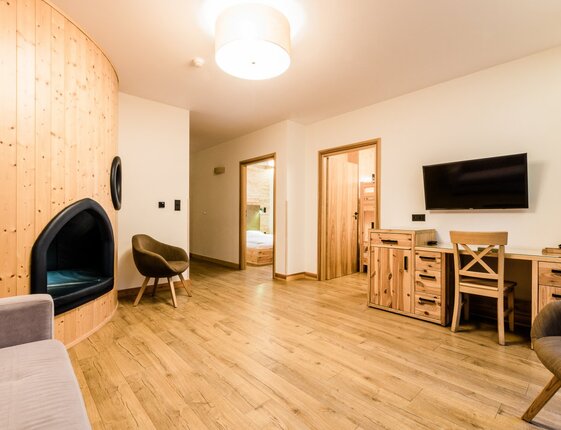 Spacious suite with curved pine seating alcove, hardwood floor, wall TV, wooden desk with kettle and chairs, doors to bedroom and wood-paneled room.