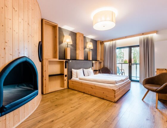 Warm wooden double room with low platform bed, bedside lamps, built-in cushioned nook, balcony with chairs and forest view.