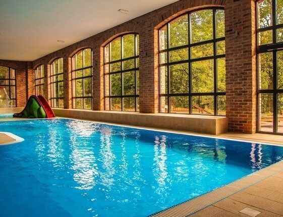 Indoor heated pool with arched brick windows, natural light, bench seating and a small children's slide overlooking trees.