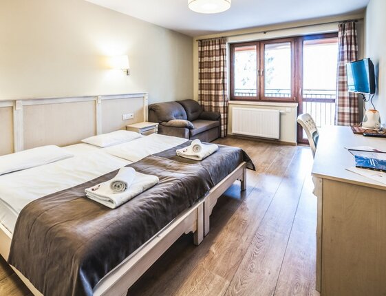 Bright twin room with two single beds pushed together, rolled towels, cozy sofa, desk with wall TV and balcony with plaid curtains